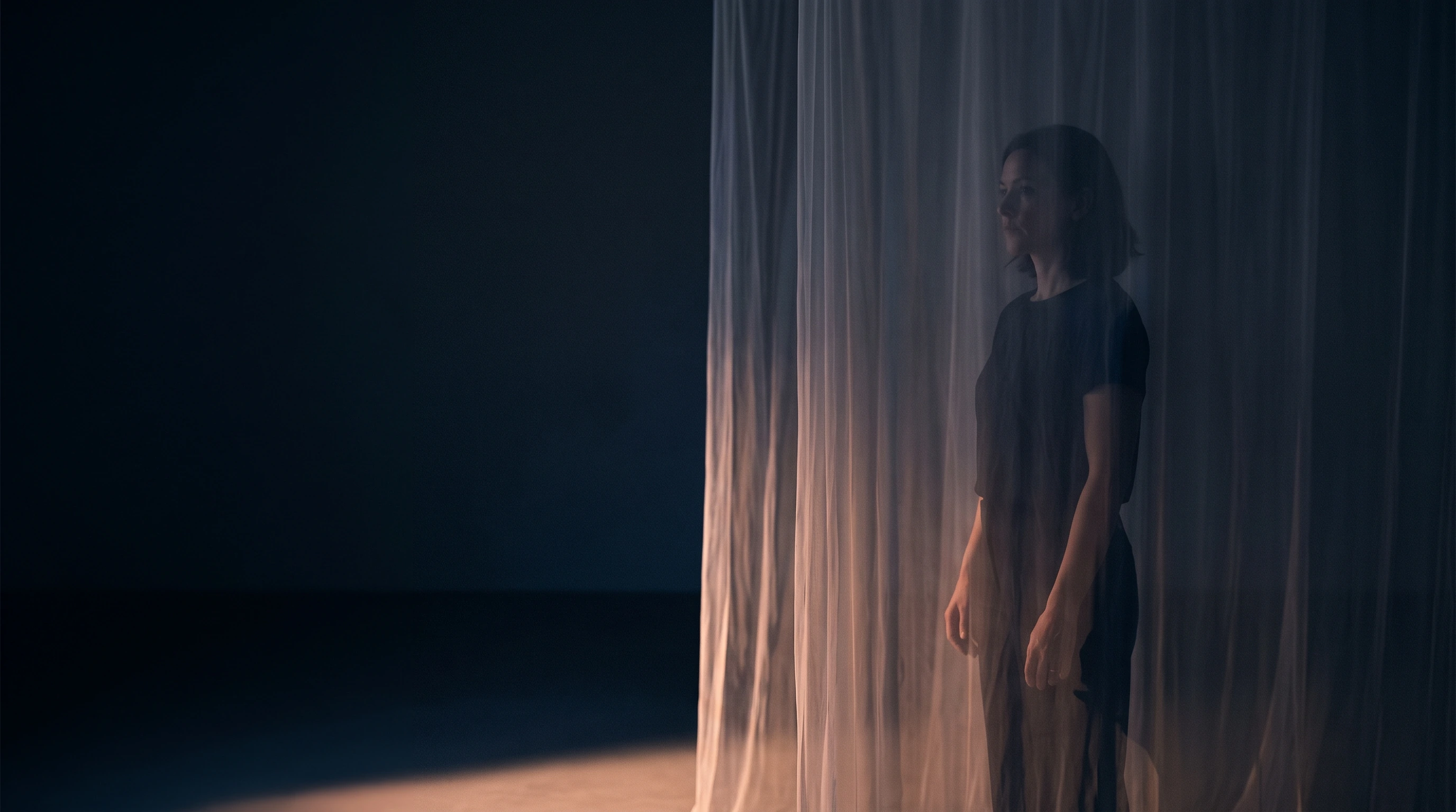 A person standing behind several layers of translucent hanging fabric, their outline clear but their face softened